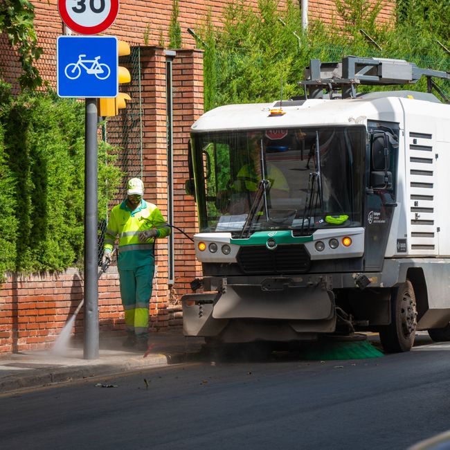 A person dressed in an orange high-visibility jacket, matching orange pants, and an orange cap is seen from above, sweeping a dusty, uneven urban sidewalk with a wide-bristle broom. They are wearing b